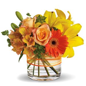 Send smiles across miles with this cheery bouquet of sunny yellow and orange blooms nestled in a compact glass vase. Featured flowers include lilies